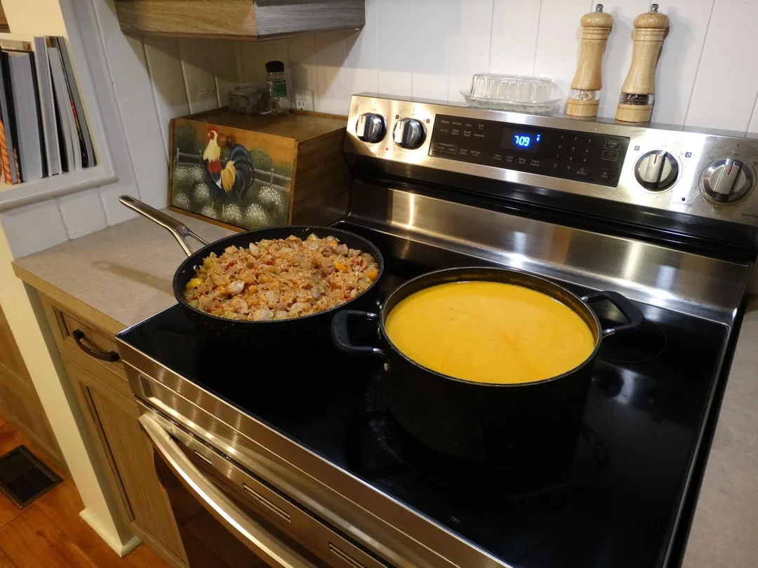 A black stovetop holds a skillet of mixed stuffing and a large pot of creamy yellow soup. Spice grinders and a rooster-themed decor sit on the counter. The oven clock reads 7:09.