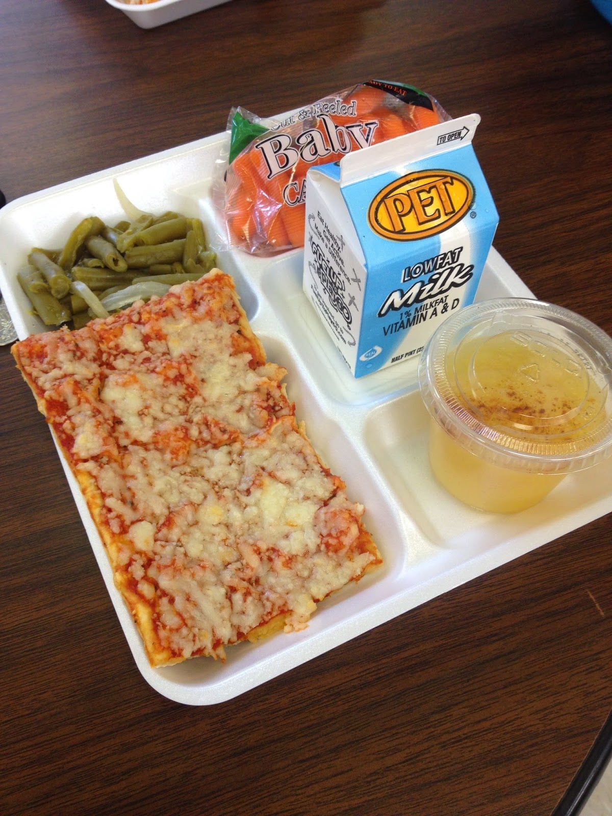 A school lunch tray with rectangular cheese pizza, green beans, a bag of baby carrots, a carton of low-fat milk, and a cup of fruit in syrup on a brown table.