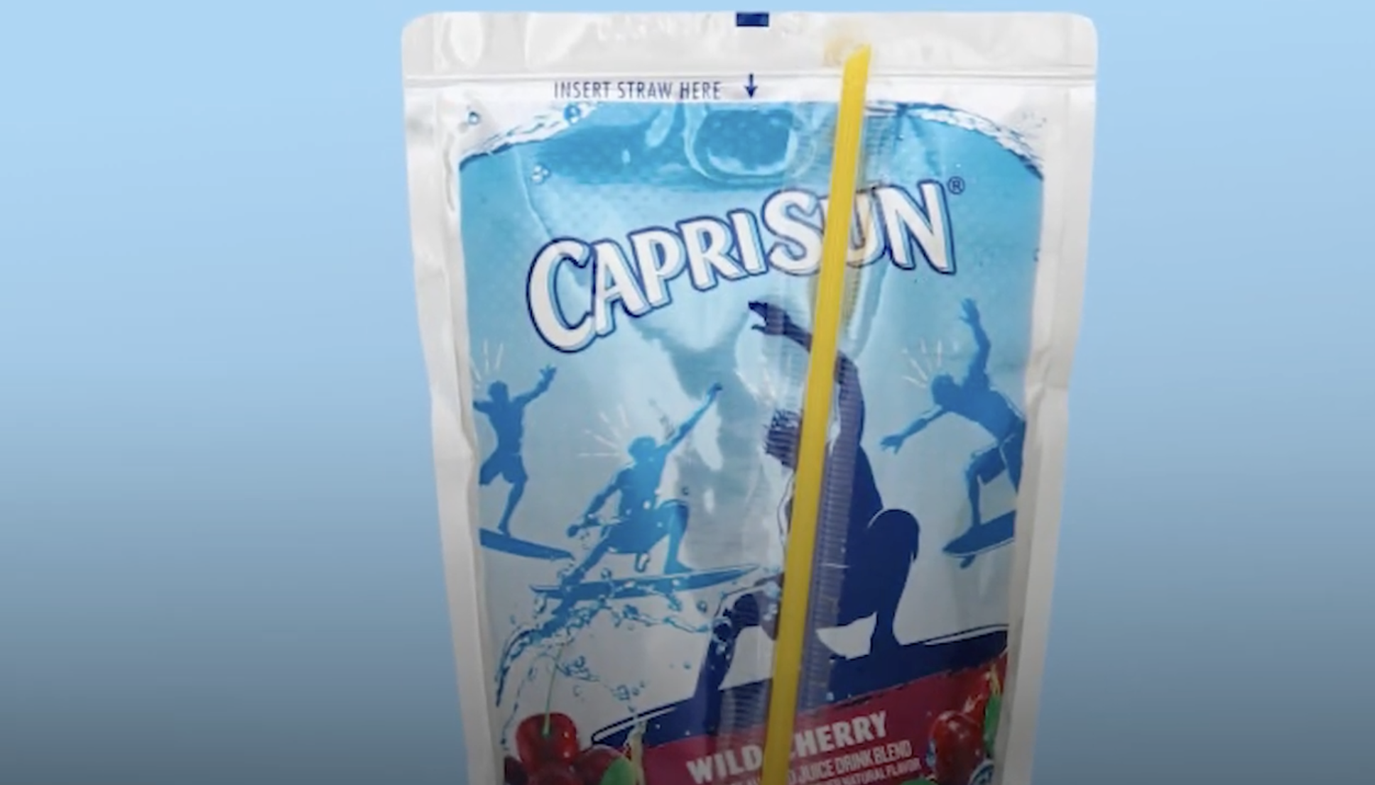 A Capri Sun juice pouch with a yellow straw inserted near the top, featuring blue graphics of surfers and labeled "Wild Cherry Juice Drink Blend" against a light blue background.