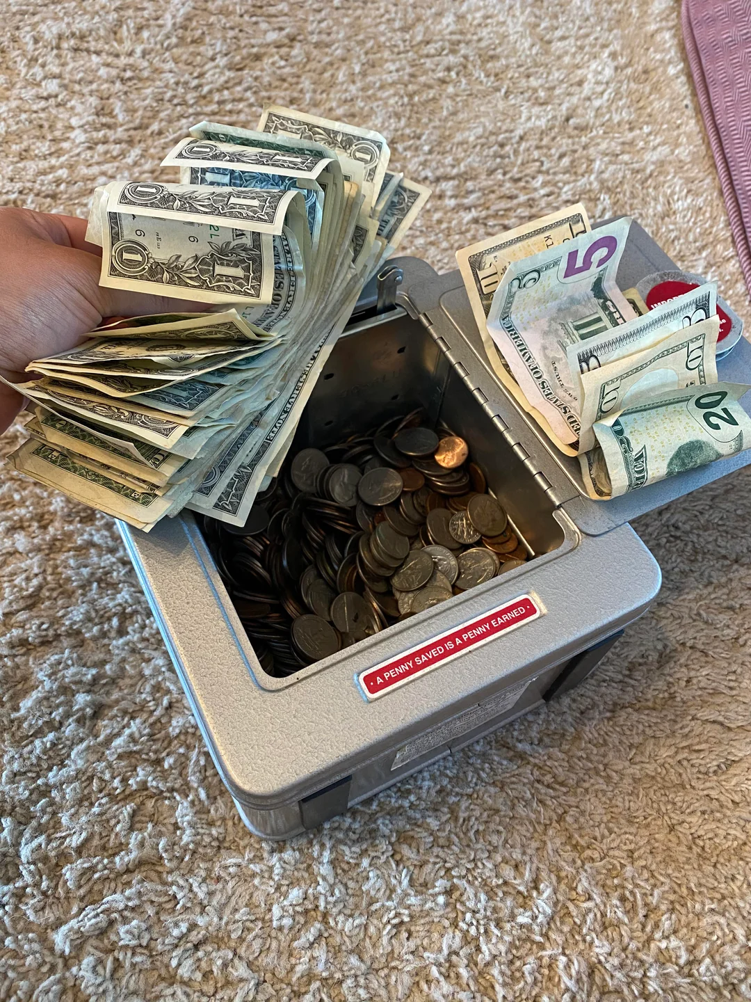 A hand holds a stack of U.S. dollar bills over an open metal cash box filled with coins and more bills, placed on a beige carpeted floor.