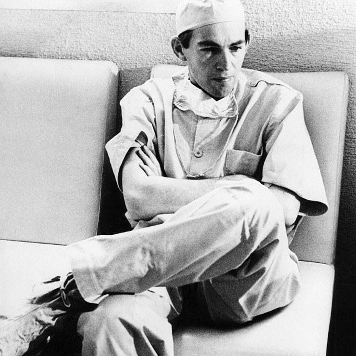 A doctor in scrubs, cap, and mask sits on a couch with arms folded, looking thoughtful. The black-and-white image gives a serious, contemplative mood.