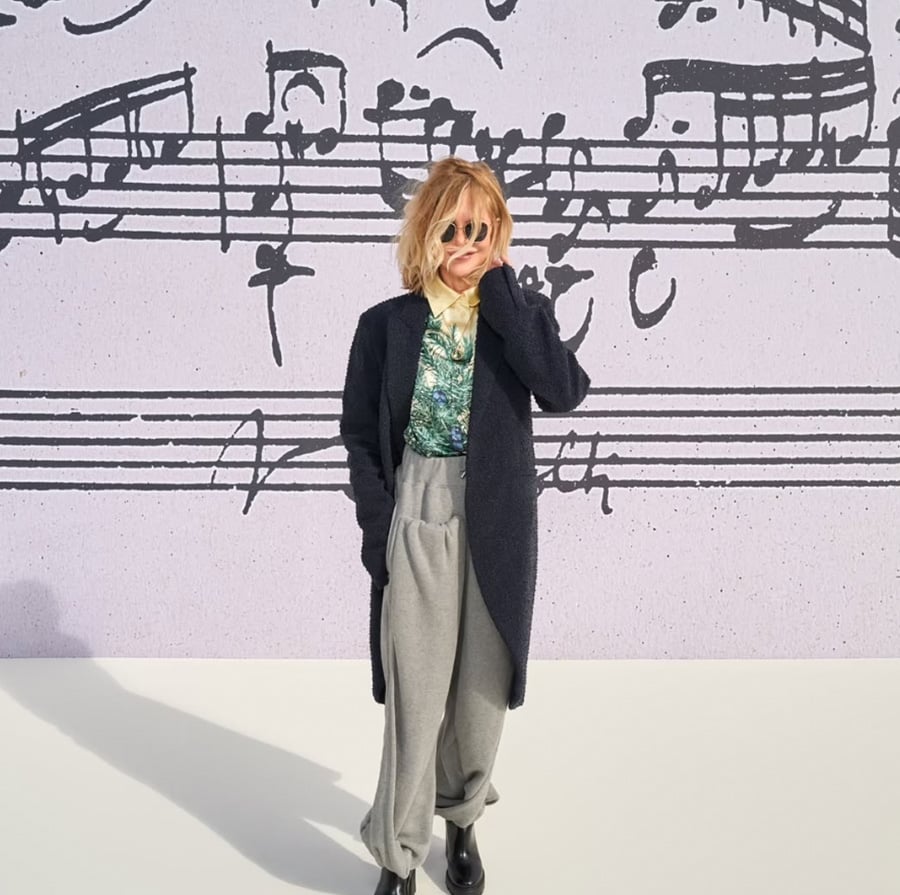 A person with shoulder-length blonde hair, sunglasses, a patterned shirt, long dark coat, and loose gray pants stands in front of a wall with large musical notes and sheet music printed on it.