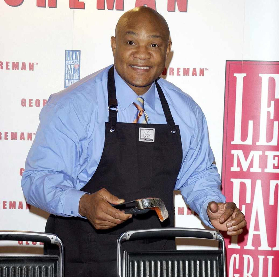 A smiling man in a blue shirt and black apron holds tongs with food, standing next to two grills. A promotional backdrop with bold red and white text is visible behind him.