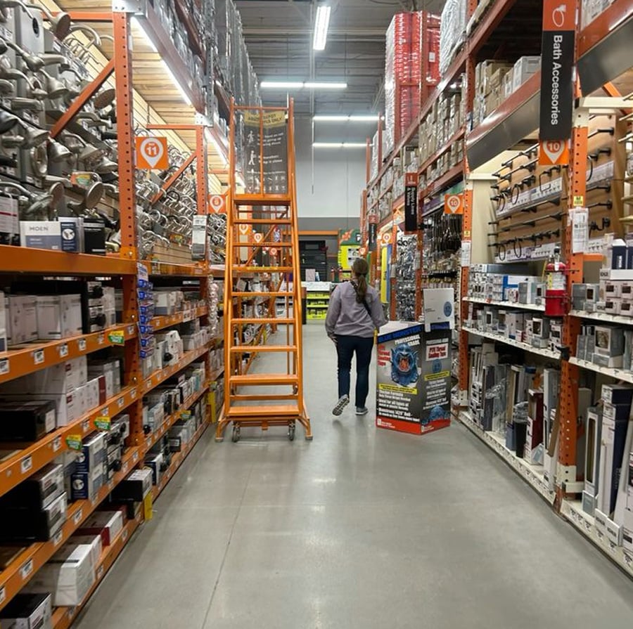 A person stands in a hardware store aisle next to an orange rolling ladder, surrounded by shelves stocked with various boxes and home improvement items. The store has high ceilings and bright lighting.