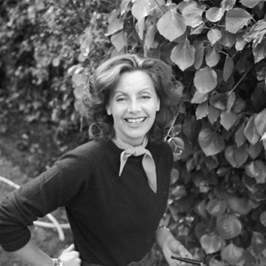 A woman with wavy hair, wearing a dark sweater and a light scarf around her neck, smiles while standing outdoors in front of leafy plants. She is holding an object in her hand.