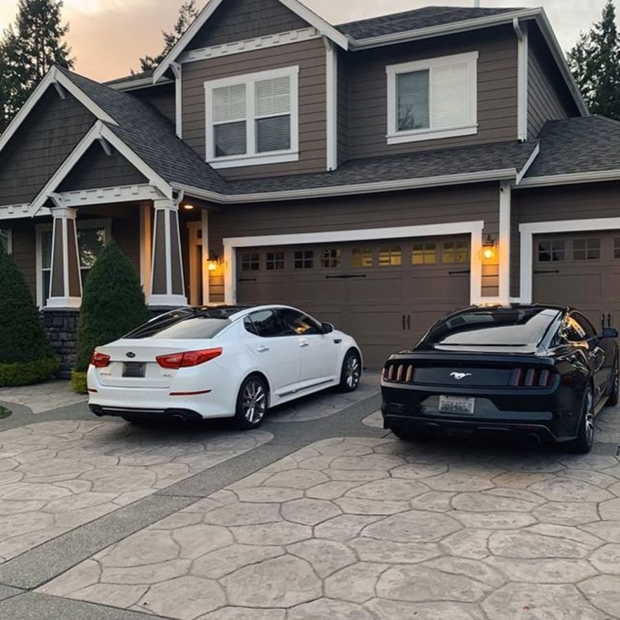 Two cars, one white and one black, are parked in the driveway of a modern two-story suburban house with brown siding and a double garage. Shrubs and trees frame the well-kept property.