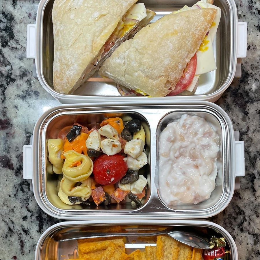 A lunchbox with three compartments: a sandwich with cheese, tomato, and meat; a pasta salad with veggies and cheese cubes; a portion of yogurt with fruit; and crackers with a small packet of spread.