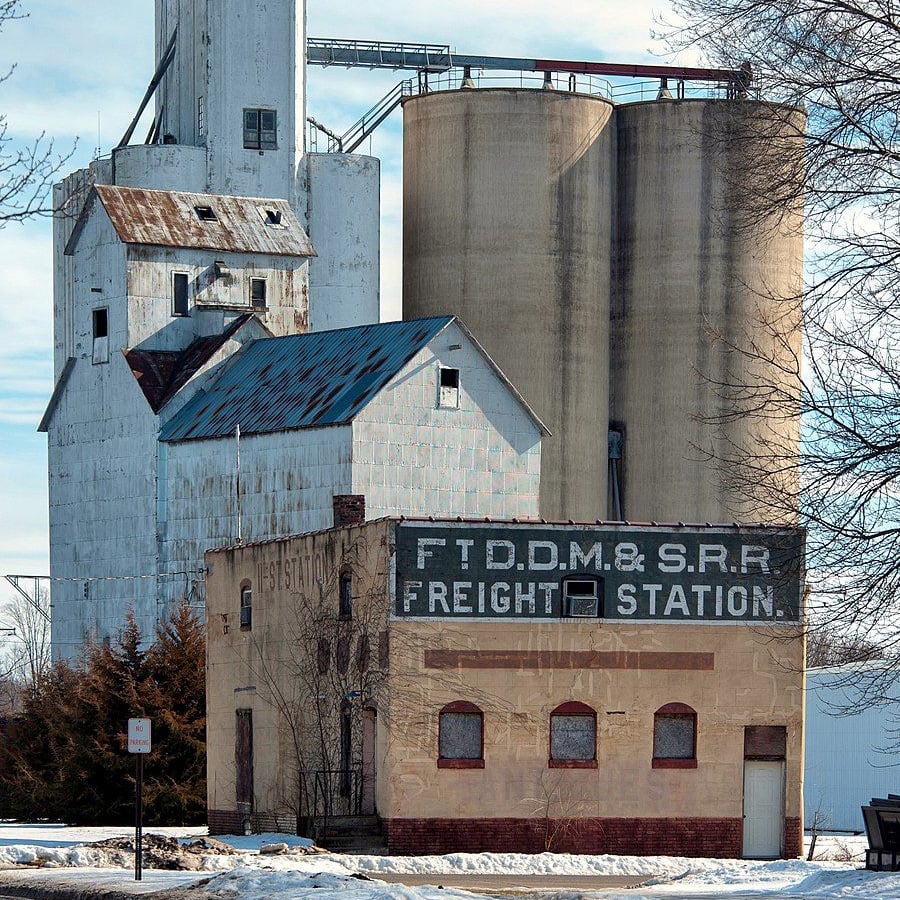A small, weathered brick building labeled "FT.D.D.M. & S.R.R. FREIGHT STATION" stands in front of tall grain silos and an old white industrial structure on a winter day, with some snow on the ground.