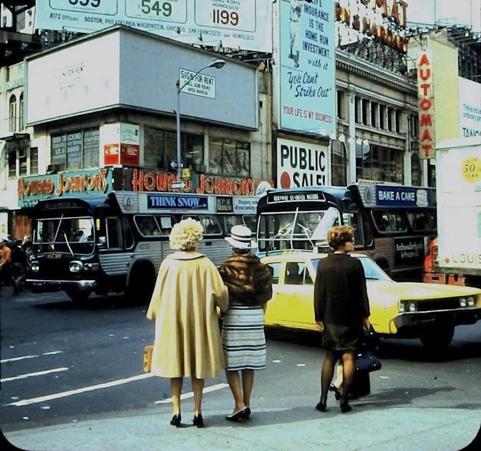 Three women cross a busy city street with vintage buses and a yellow car nearby. Billboards, signs, and storefronts, including “Howard Johnson’s” and “AUTOMAT,” are visible in the background.