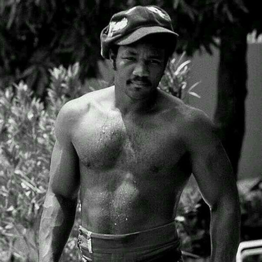 A muscular man with a mustache stands shirtless outdoors, wearing a cap and pants. Trees and foliage are visible in the background. The image is in black and white.