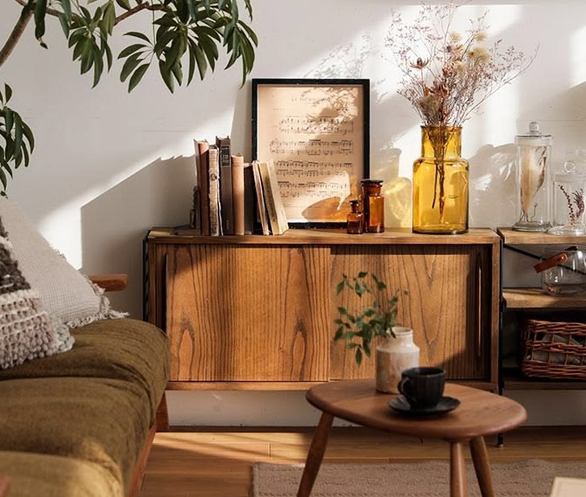 A cozy living room corner with a wooden cabinet, a framed sheet of music, dried flowers in amber vases, books, a small plant, and a round table with a cup, all bathed in warm natural sunlight.