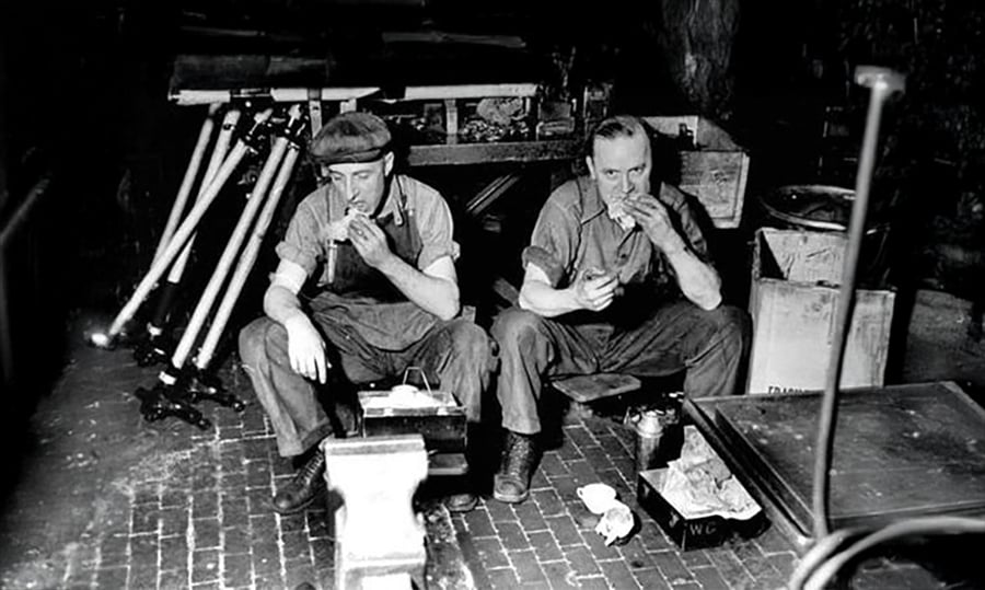 Two men in work clothes sit on low stools in an industrial workshop, taking a lunch break and eating sandwiches. Various tools, equipment, and boxes surround them on a brick floor.