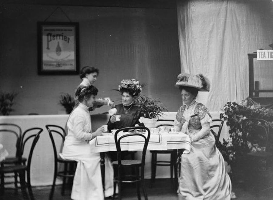 Three women in Edwardian-era dresses and hats sit at a table enjoying tea, while another woman stands pouring tea. The setting appears to be a vintage tea room with simple decor and a Perrier sign on the wall.