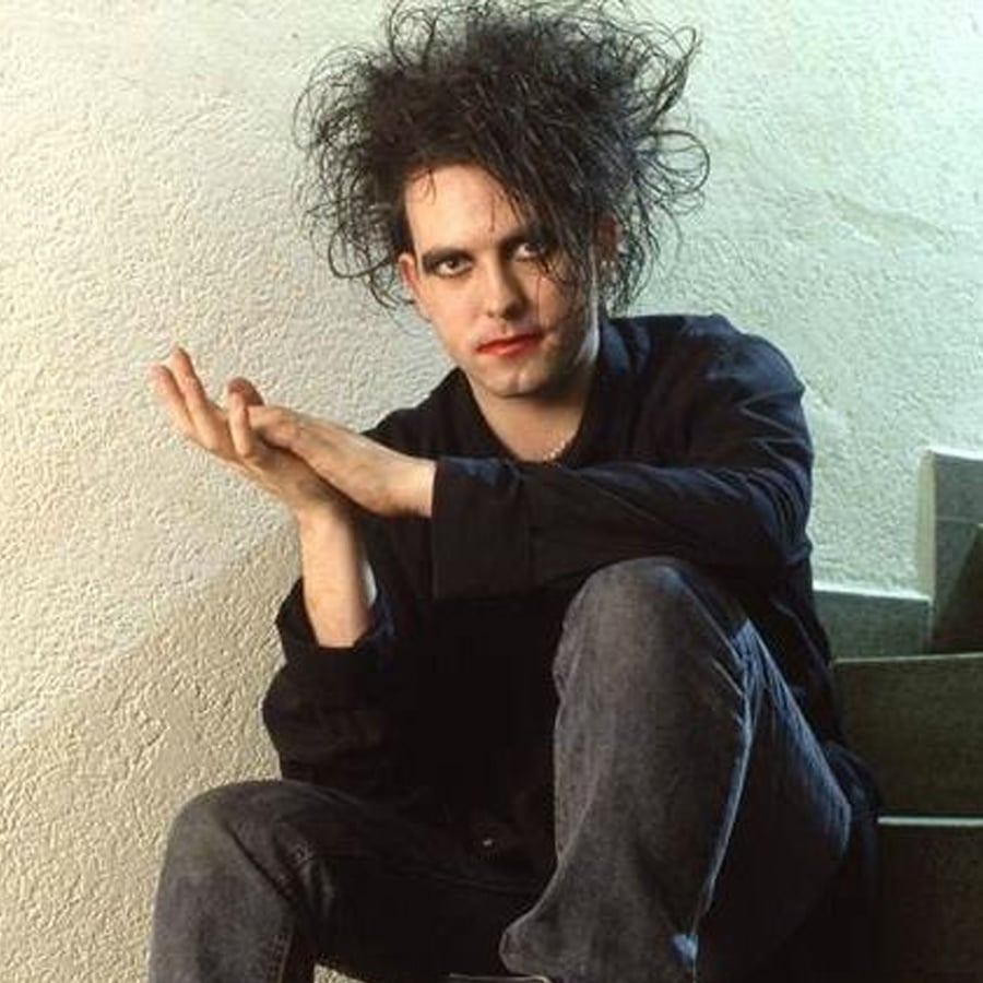 A person with pale skin, heavy black eye makeup, and messy black hair sits on stairs against a white wall, wearing a loose black shirt and dark jeans, hands together in front, looking at the camera with a neutral expression.