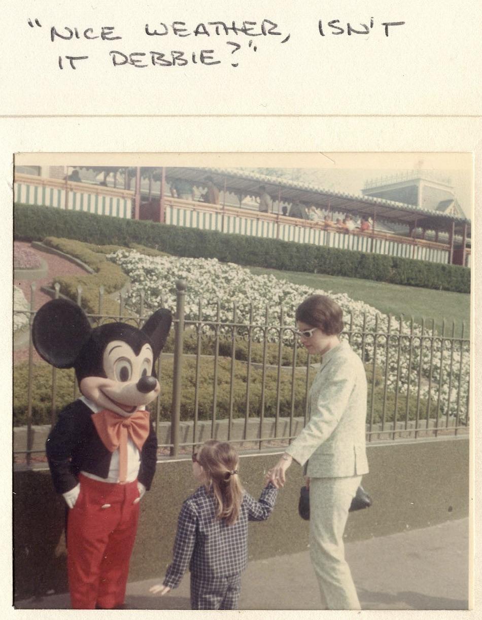 A child holding hands with an adult stands near a person in a Mickey Mouse costume at an amusement park. The caption above reads, "Nice weather, isn't it Debbie?" in handwritten text.