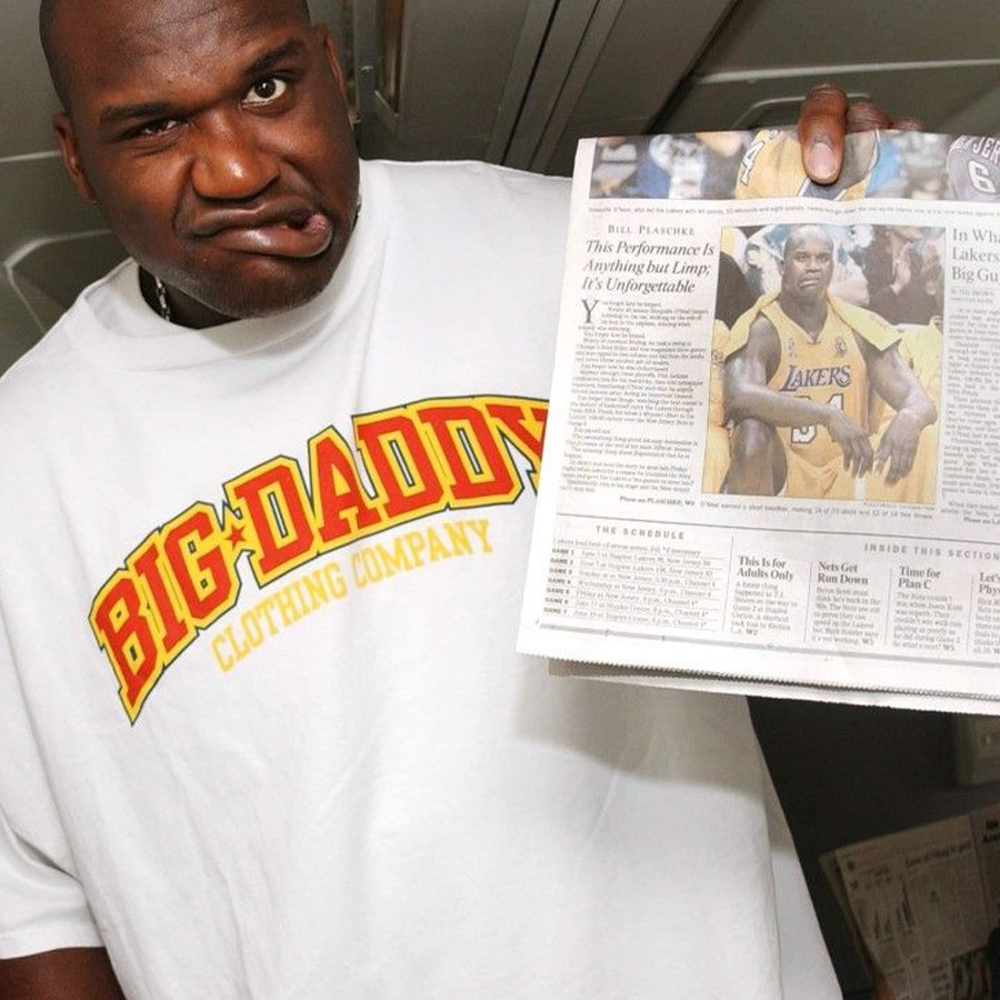 A man in a "Big Daddy Clothing Company" shirt makes a funny face while holding up a newspaper featuring a photo of him playing basketball for the Lakers.