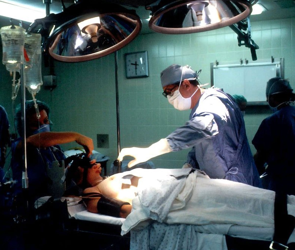 A medical team performs surgery in an operating room. A patient lies on the table under bright surgical lights, surrounded by doctors in scrubs, masks, and gloves. IV bags hang nearby, and medical equipment is visible.
