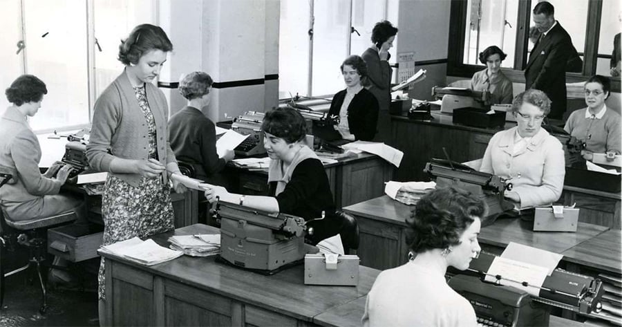 Black and white photo of women working at desks with typewriters in an office, while one woman stands handing papers to a seated colleague. A man stands in the background, observing the busy workspace.