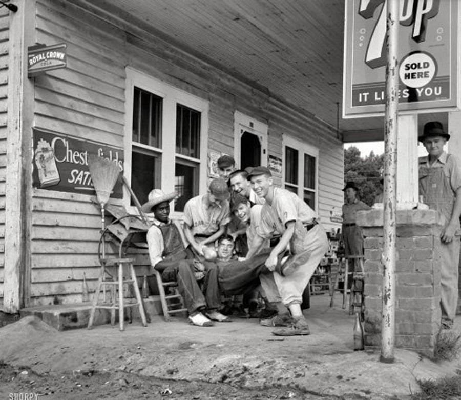 A group of men and boys gather playfully on the porch of an old wooden store with vintage signs for 7-Up, Chesterfields, and Royal Crown Cola. Some sit and some stand, while one leans against a post and another looks on.