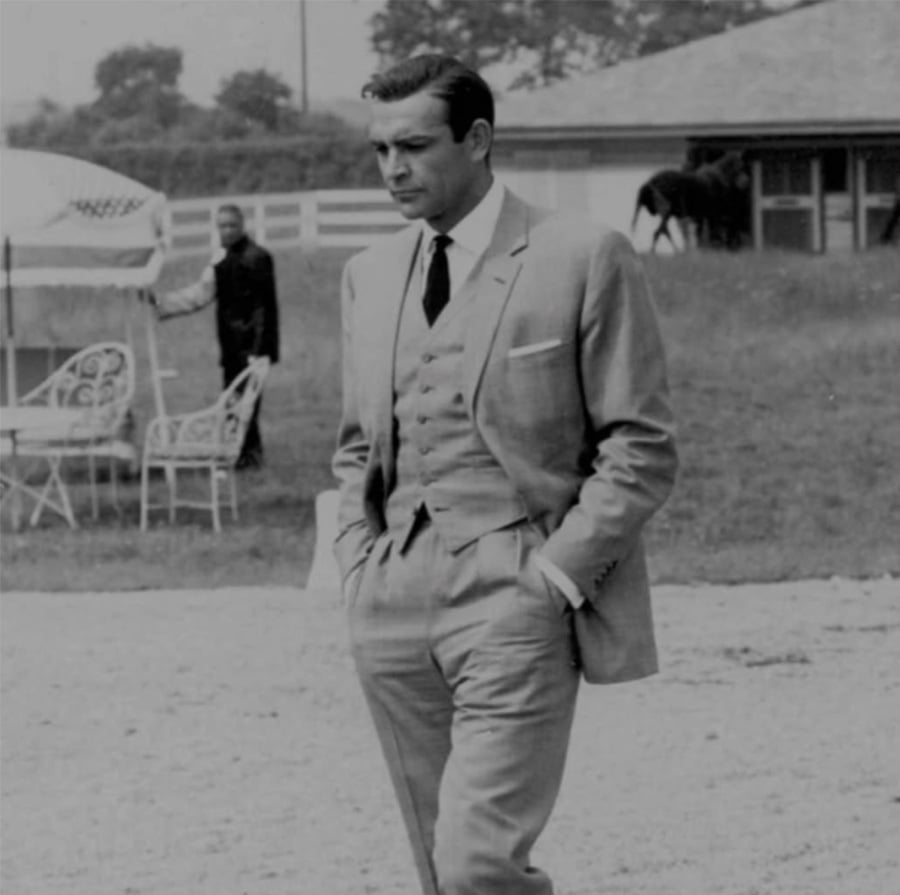 A man in a light-colored suit walks outdoors with his hands in his pockets. There is outdoor furniture, another person in the background, and a horse near a building. The image is in black and white.
