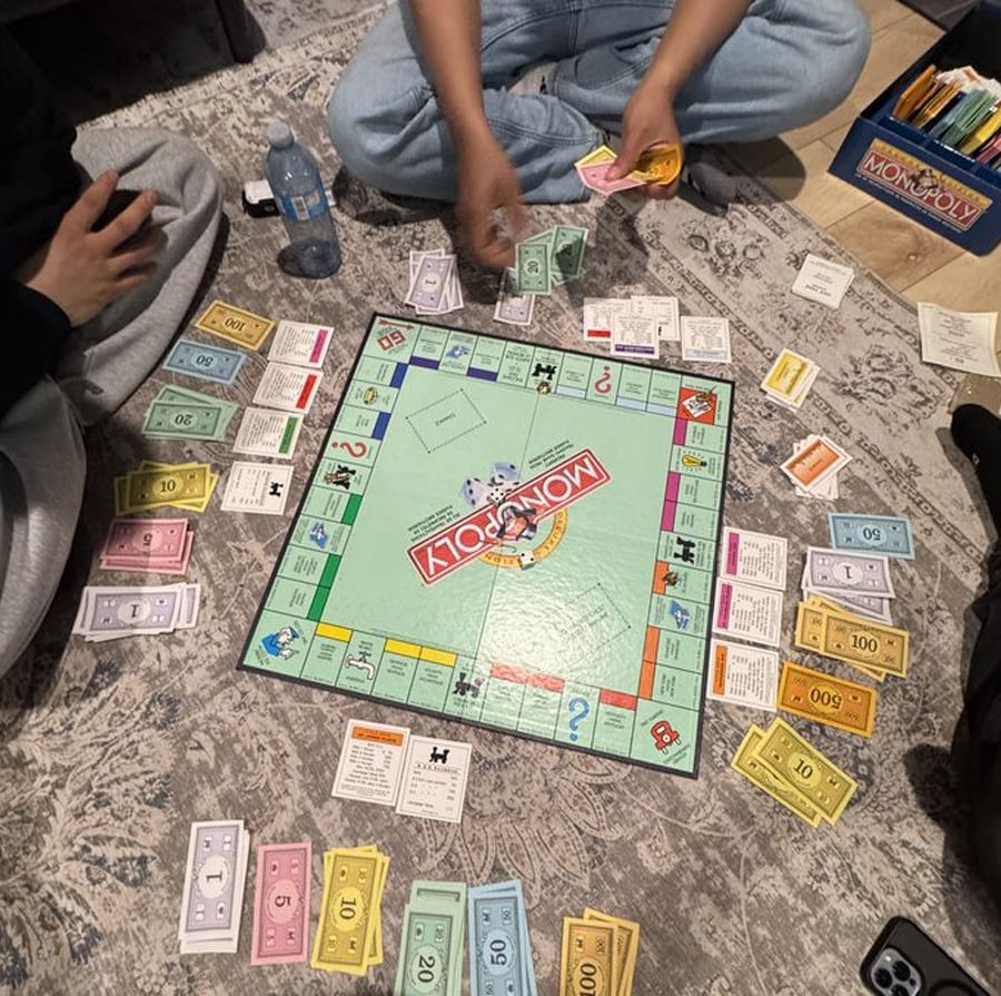 Three people are playing Monopoly on a patterned carpet. Monopoly money, property cards, and playing pieces are spread around the board, with hands visible handling cash. The Monopoly box and a water bottle are nearby.