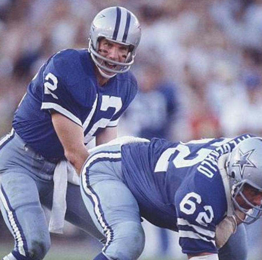 Two Dallas Cowboys football players in blue uniforms are on the field; the player wearing number 12 is positioned behind the center, number 62, preparing to receive the snap during a game.