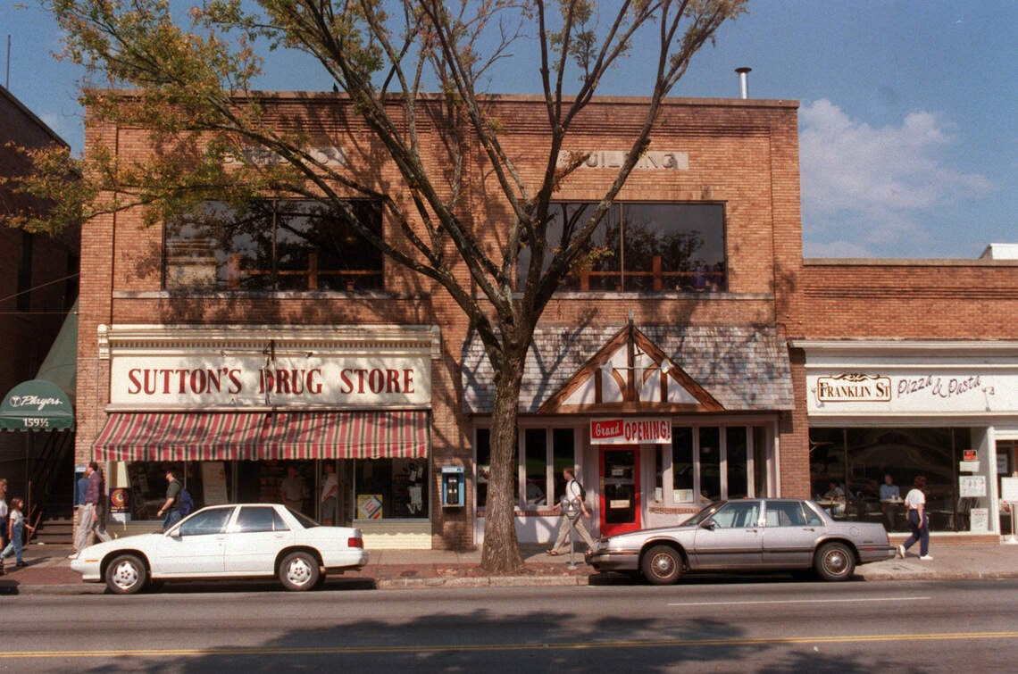 A brick building houses Sutton’s Drug Store, a restaurant, and Franklin’s Street Pizza & Pasta. Two cars are parked on the street and people are walking on the sidewalk under a large tree.