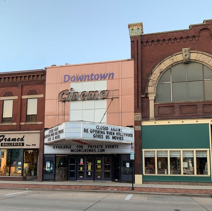A vintage Downtown Cinema with a marquee sign that reads, "CLOSED AGAIN: REOPENING WHEN HOLLYWOOD GIVES US MOVIES." The theater is flanked by a gallery and a storefront on a quiet street.
