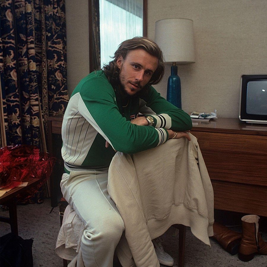 A man with shoulder-length hair and a beard wears a green and white tracksuit, sitting on a chair and leaning forward over a jacket. Behind him are a desk, lamp, TV, and a pair of brown boots on the floor.