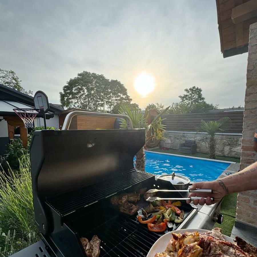 A person grills meat and vegetables on a barbecue next to a swimming pool at sunset. A basketball hoop, greenery, and houses are visible in the background.