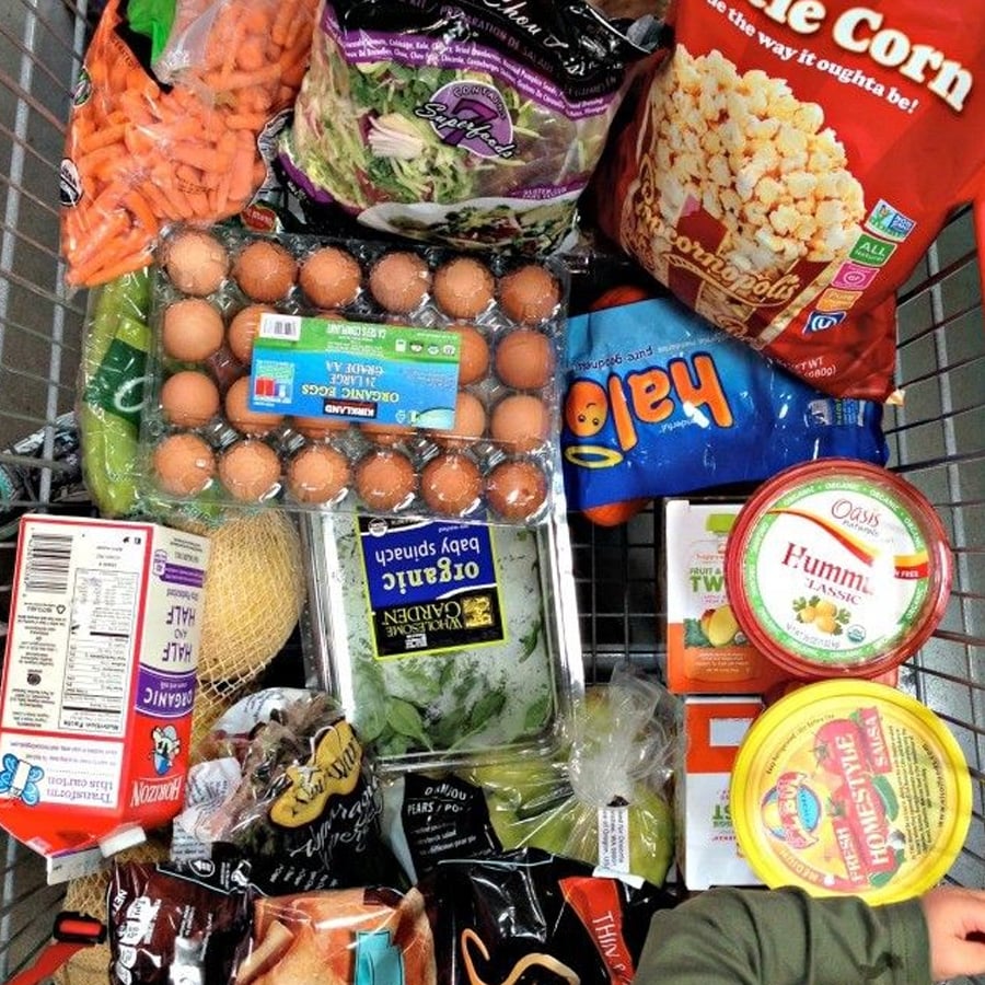 A grocery cart filled with various items including eggs, salad mix, baby spinach, hummus, popcorn, Halo mandarins, milk, carrots, bananas, and other packaged foods.