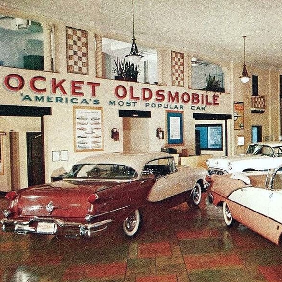 Classic 1950s Oldsmobile cars are displayed inside a vintage dealership with a large sign reading "ROCKET OLDSMOBILE 'America's Most Popular Car'" on the wall above them.