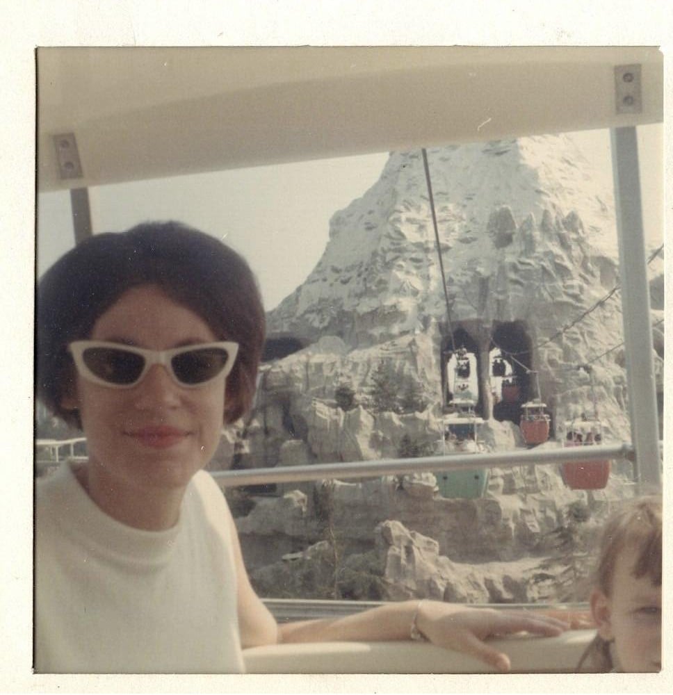 A woman in cat-eye sunglasses smiles while sitting on a ride in front of Disneyland’s Matterhorn mountain, with cable cars and rocky scenery visible in the background.