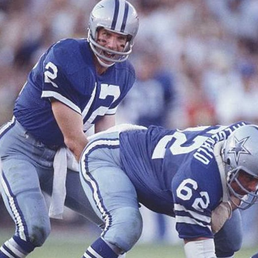 Dallas Cowboys quarterback in a blue uniform prepares to receive the snap from the center during a football game, with both players focused and ready to start the play.