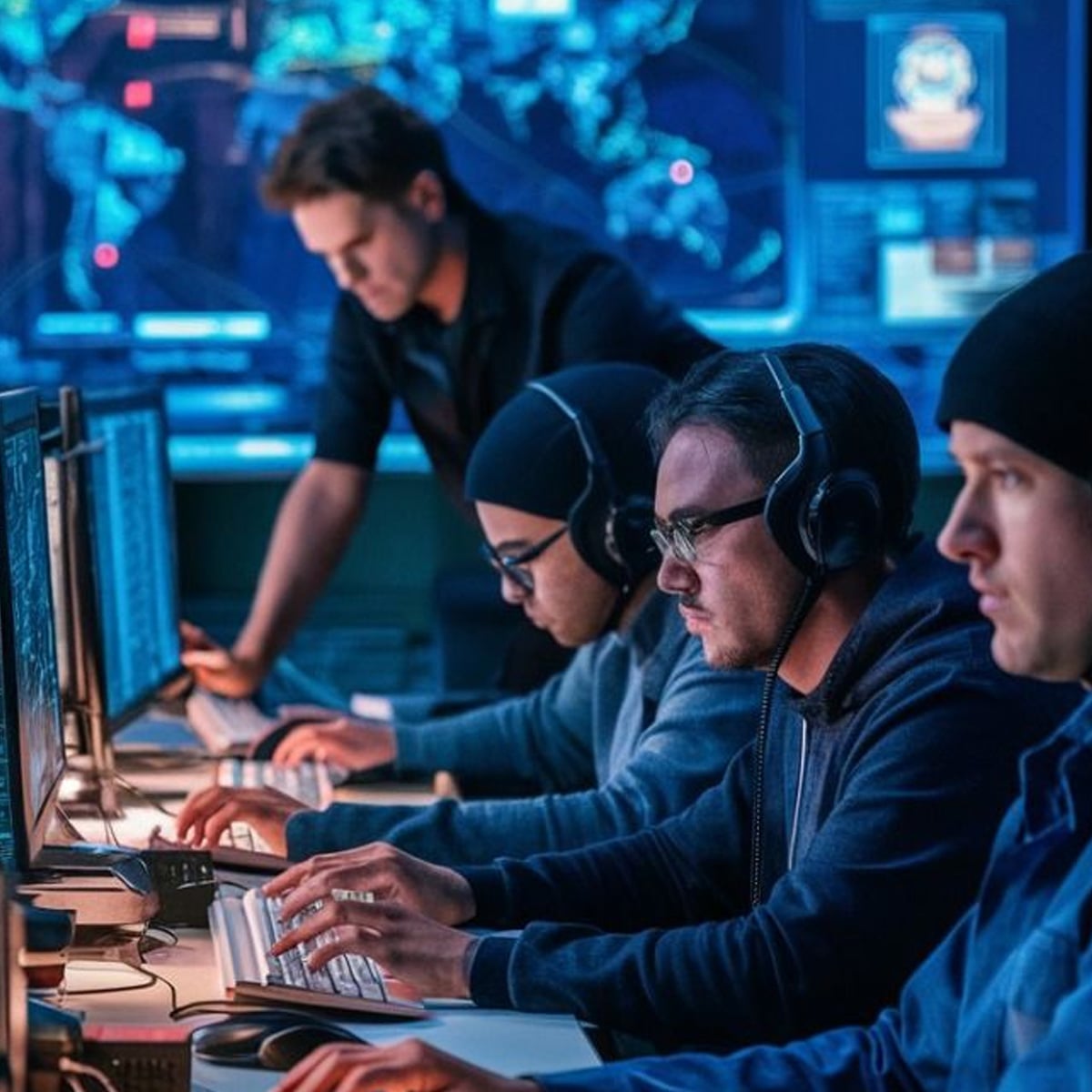 Four people wearing headsets and dark clothing work intently at computer monitors in a dimly lit room with large screens showing maps and data in the background. One person stands behind them, supervising.