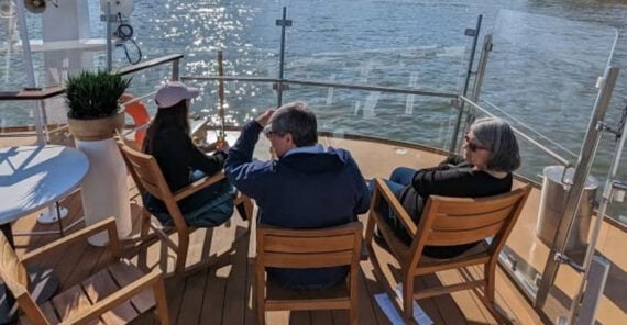 Three people sit on wooden chairs on a boat deck, facing the water on a sunny day. The water sparkles in the sunlight, and the group appears to be relaxing and enjoying the view.