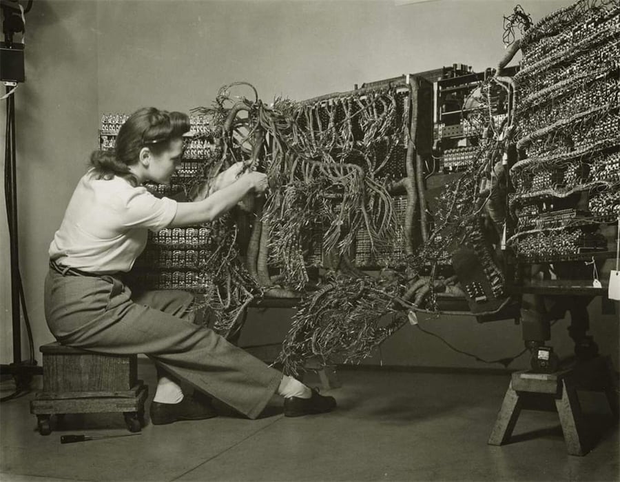 A woman sits on a stool, working with a complex array of cables and wires on an early computer system, likely from the mid-20th century, in a black and white photograph.
