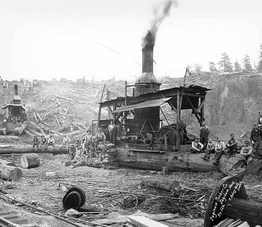 A group of workers sit and stand on and around a large steam-powered logging machine in a deforested area, surrounded by fallen logs and debris, with smoke rising from the machine’s smokestack.