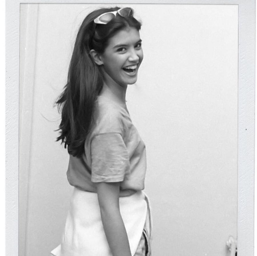 A young woman with long hair smiles brightly, looking over her shoulder. She wears a casual t-shirt, has sunglasses on her head, and a sweater tied around her waist. The photo is black and white with a simple background.