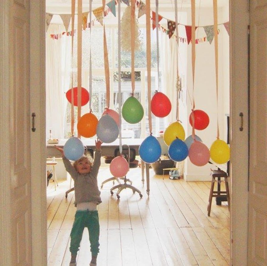 A young child reaches up toward colorful balloons that are hanging from the ceiling with ribbons in a bright, decorated room with wooden floors and festive bunting.