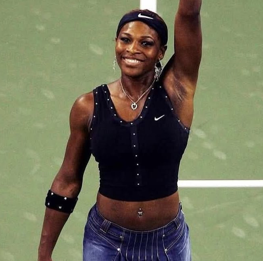 A female tennis player wearing a black sleeveless top, denim skirt, and headband smiles and raises her arm on a tennis court.