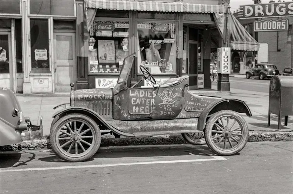 A vintage open-top car is parked on a city street. The car has handwritten messages on the side, including “NO LADIES ENTER HERE!” Behind it are storefronts, including a drugstore and a soda shop.