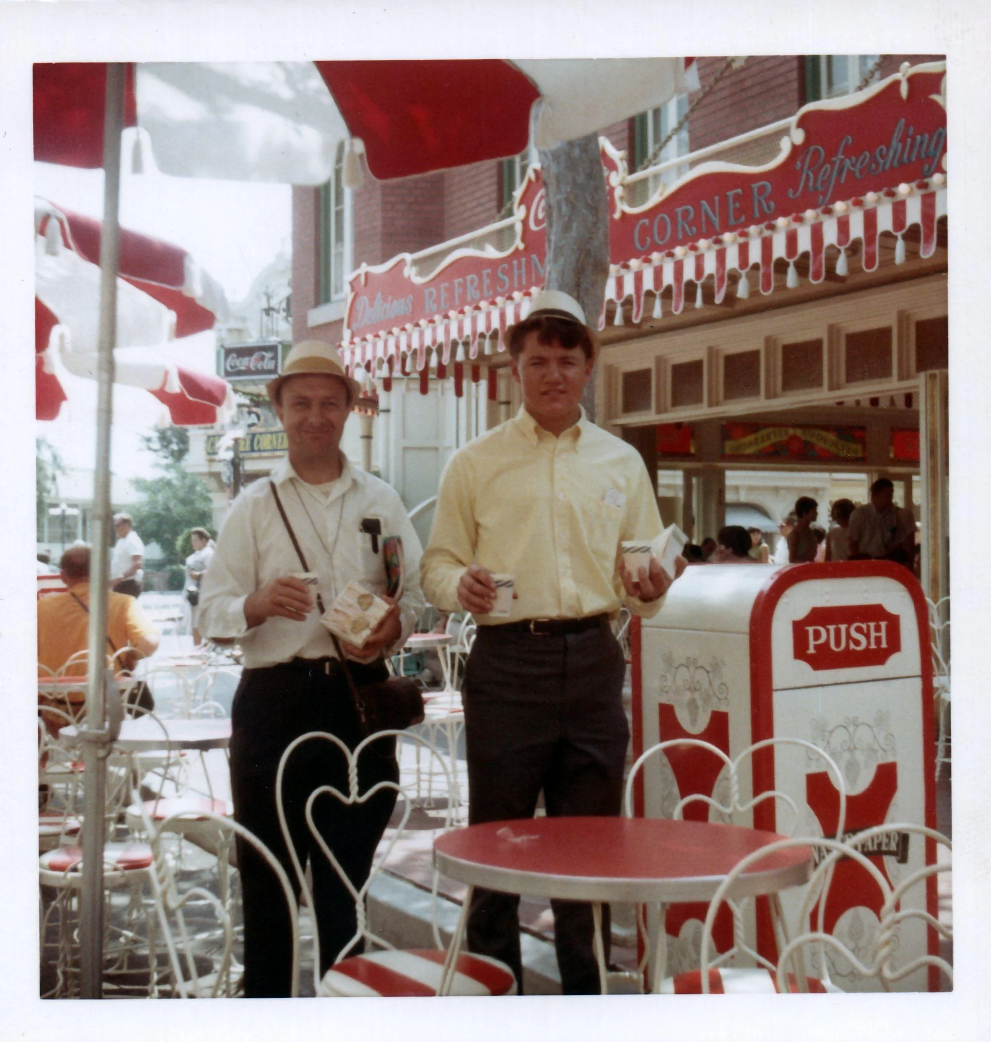 Two men stand outside a vintage soda shop holding snacks, under red and white umbrella tables. The shop’s sign reads “Refreshing Corner,” and people are seated at patio tables nearby. A Coca-Cola sign is visible in the background.