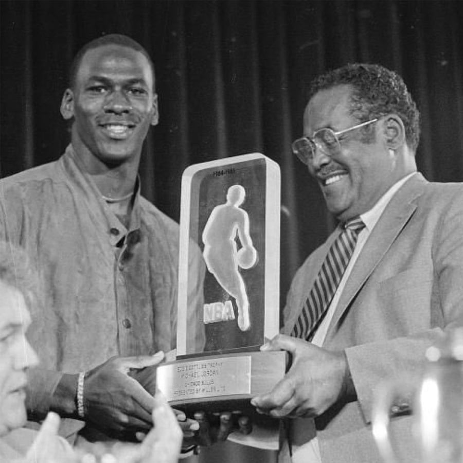 A smiling basketball player receives an NBA trophy from a man in a suit during an award ceremony. Both appear happy, and the trophy has a silhouette of a player and reads "NBA.