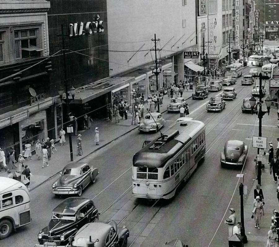 Black and white photo of a busy city street from the mid-20th century, featuring a streetcar, vintage cars, pedestrians, and storefronts, including Macy’s and Allen’s, with visible streetcar tracks.