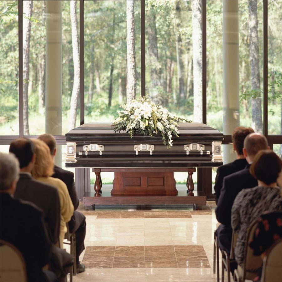A group of people sit facing a closed casket adorned with white flowers in a bright room with large windows overlooking trees, suggesting a funeral or memorial service.