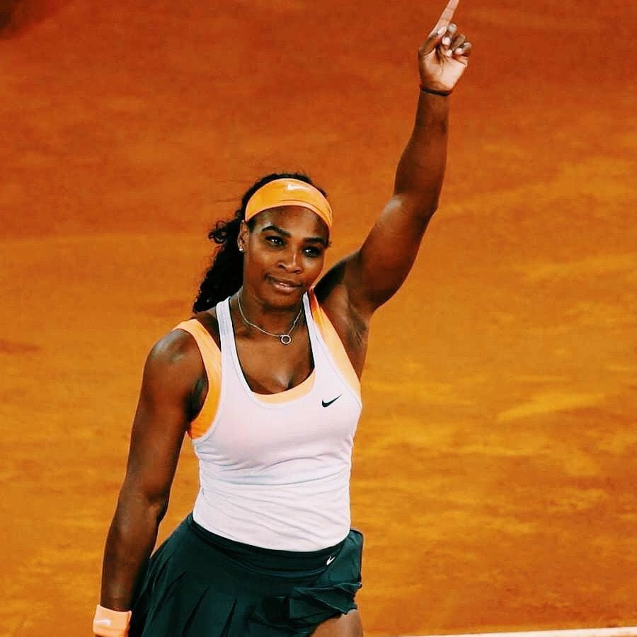 A female tennis player on a clay court raises her right arm and points upward, wearing a white and orange top, dark skirt, orange wristband, and a matching headband. She appears confident and focused.