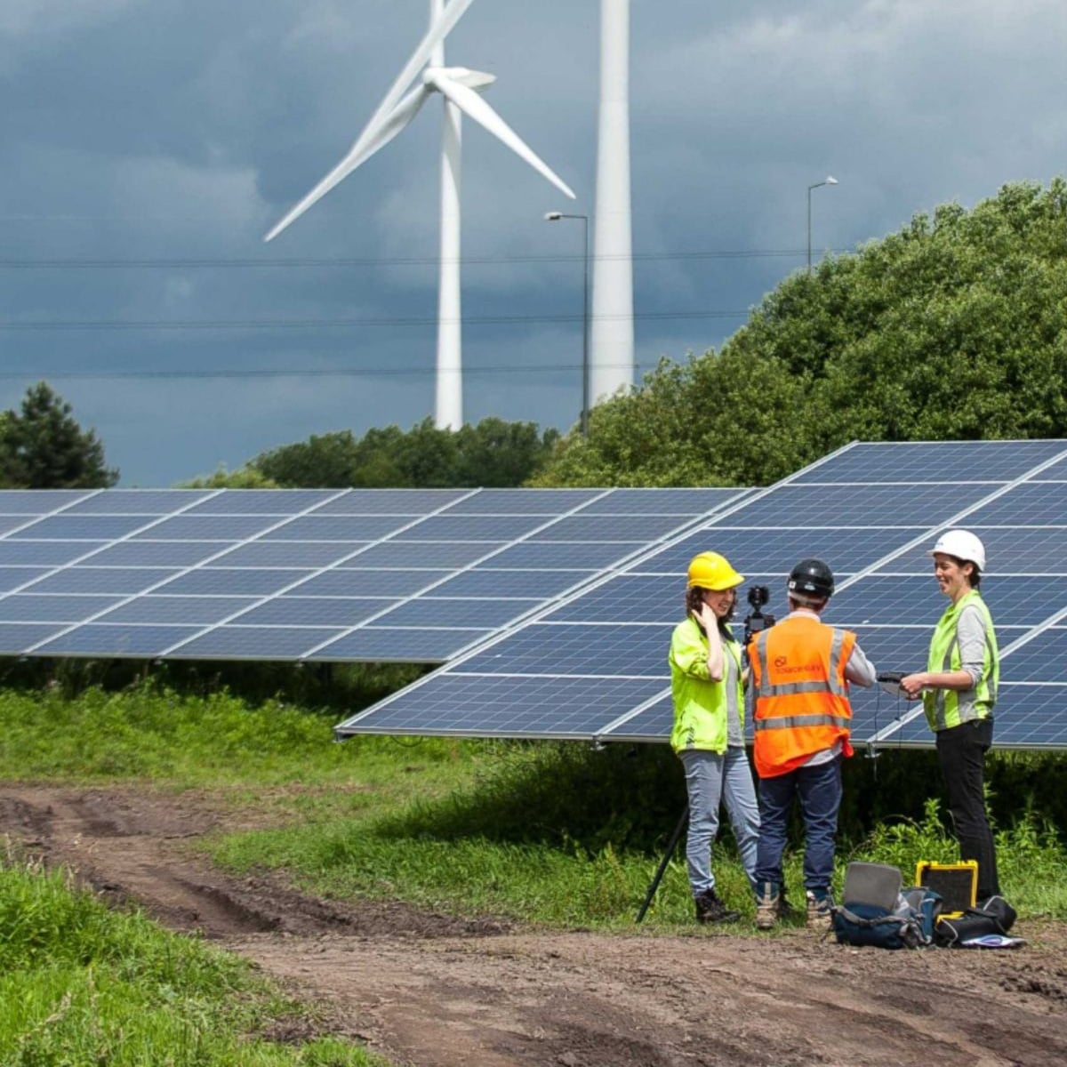 Three people wearing safety gear stand in front of solar panels in a grassy area, discussing or examining equipment, with two large wind turbines visible in the background under a cloudy sky.
