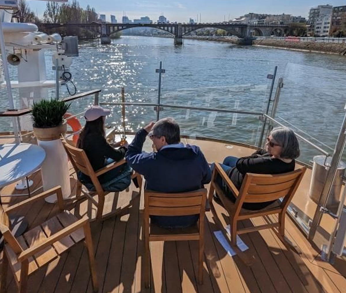 Three people sit on wooden chairs at the back of a boat, facing a river and a distant bridge on a sunny day, with city buildings and trees visible along the riverbanks.