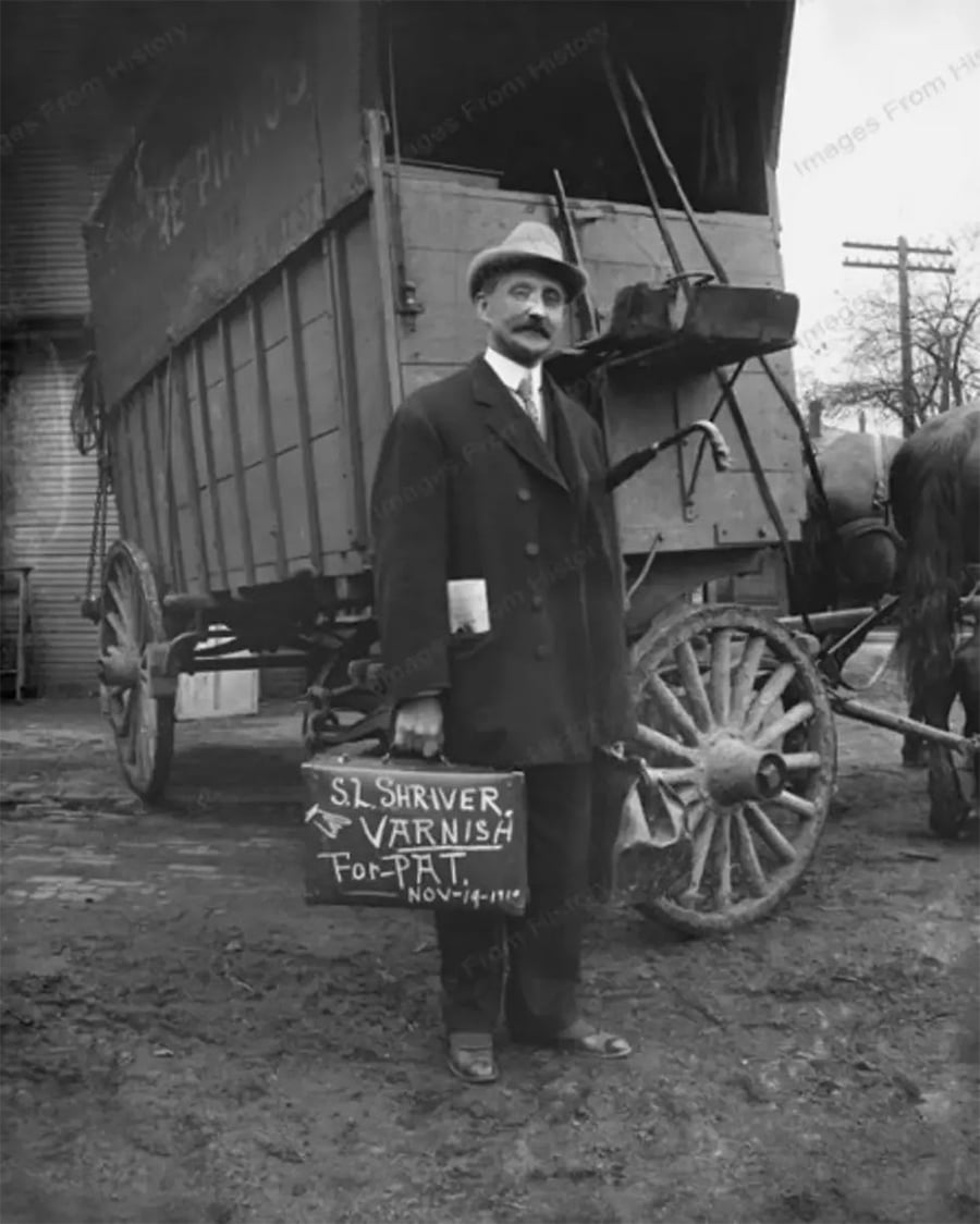 A man in a suit and hat stands in front of a horse-drawn wagon, holding a briefcase labeled “S.L. Shriver Varnish for Platt, Nov. 18, 1910.” The ground is muddy and a horse is partially visible on the right.